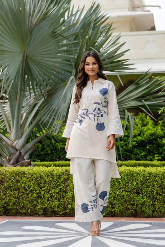 Woman in a cream outfit with blue floral patterns standing in front of tropical plants.
